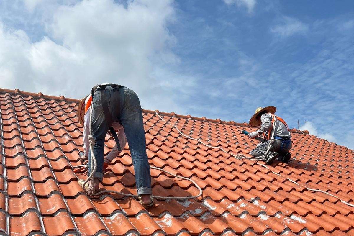 Two workers wearing hats and harnesses repairing a red tiled roof under a blue sky.