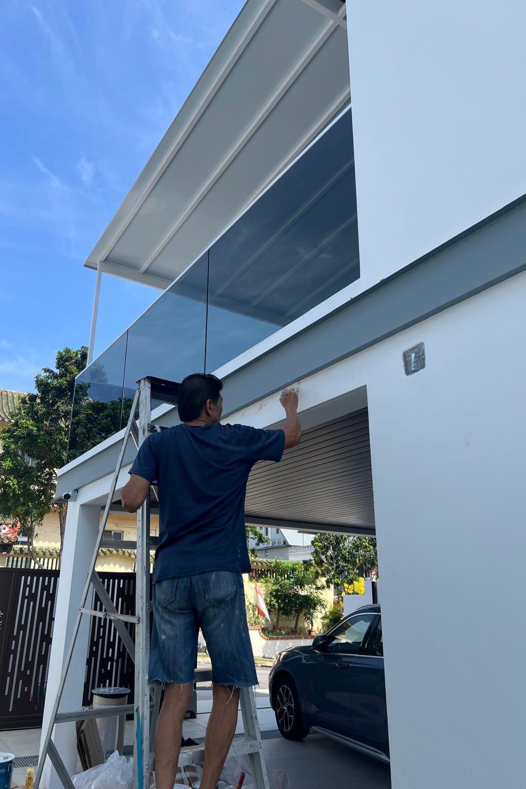 Man on ladder painting the white exterior wall of a modern house with glass balcony and parked car nearby.