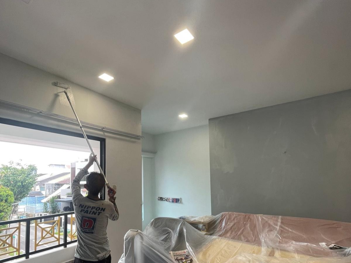 Painter painting a white wall near a window in a modern living room with plastic-covered furniture.