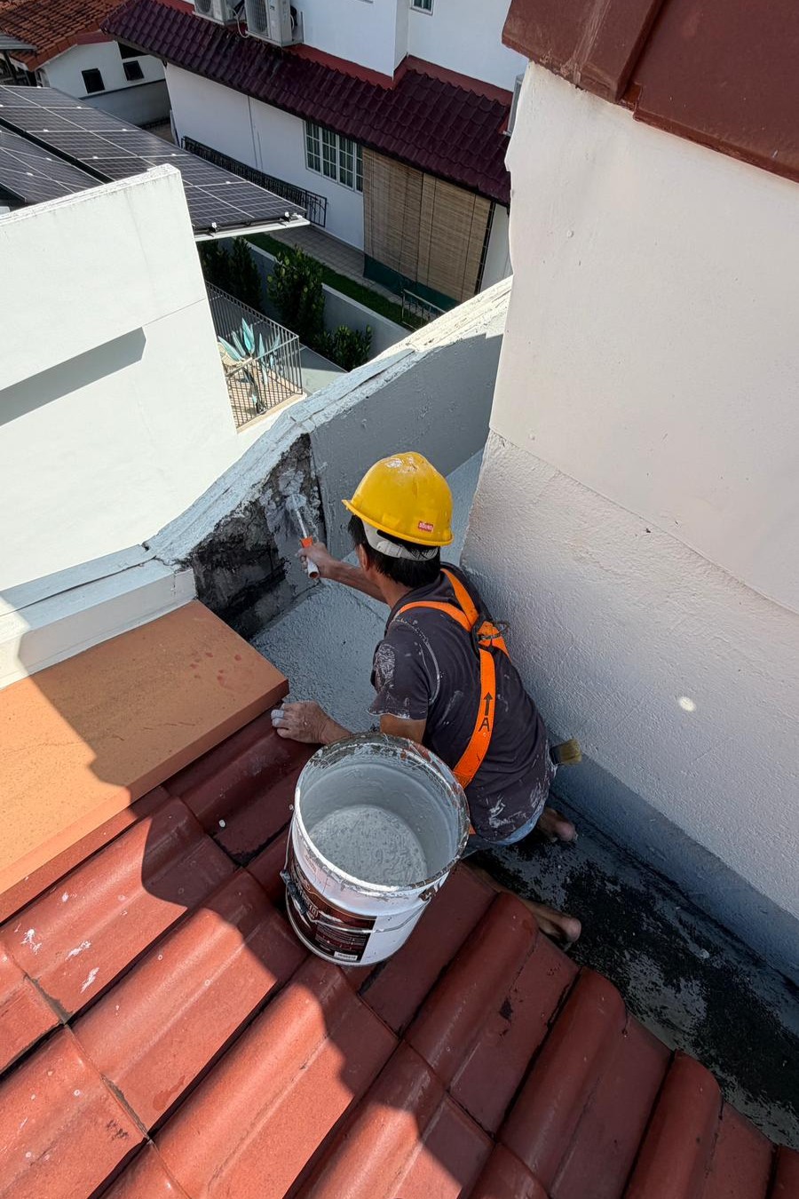 Worker in yellow helmet painting a white corner wall on a rooftop with red tiles and a bucket of paint.