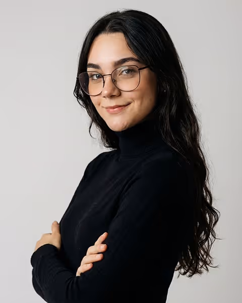 Woman with long dark hair and glasses wearing a black turtleneck smiling with arms crossed.