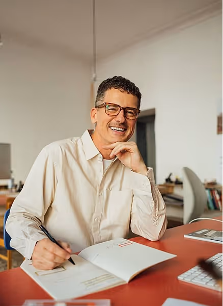 Man wearing glasses smiling and writing in an open notebook at a desk in a home office.