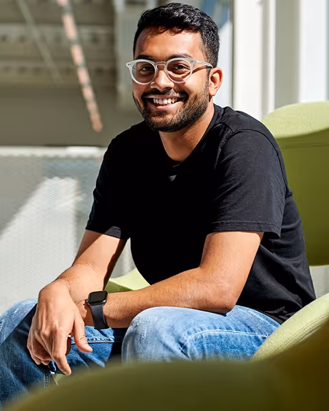 Smiling man with glasses wearing black t-shirt and jeans sitting on a green chair in bright room.