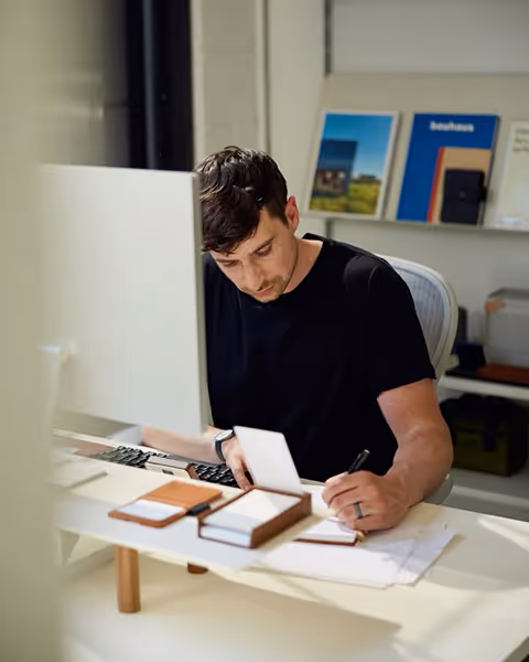 Man in a black shirt writing on paper at a desk with a computer and organized stationery.