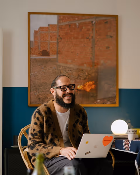 Smiling man with glasses and beard using laptop while sitting on a chair in a cozy room.