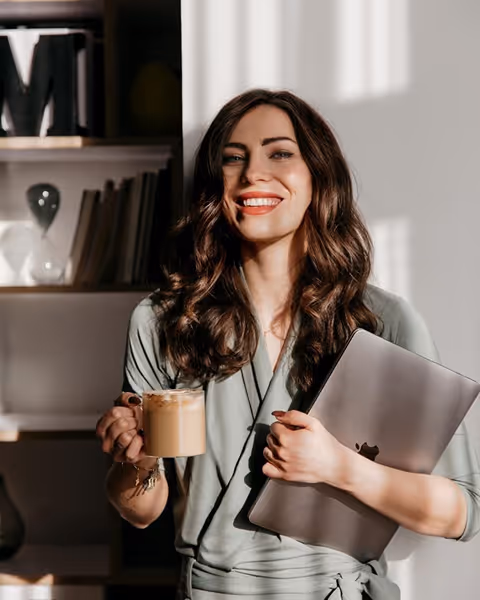 Smiling woman holding a coffee mug in one hand and a closed laptop in the other indoors.