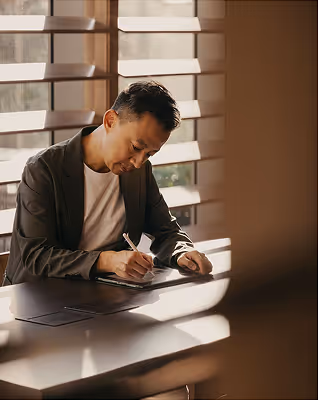 Man sitting at a table by a window writing on a clipboard in soft sunlight.