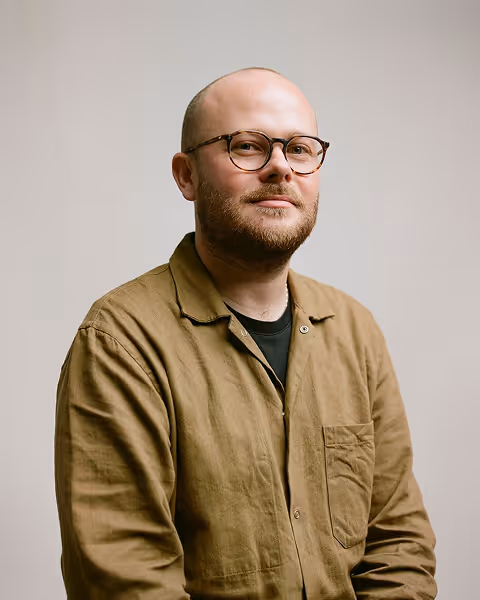 Man with a beard and glasses wearing a brown button-up shirt against a plain background.