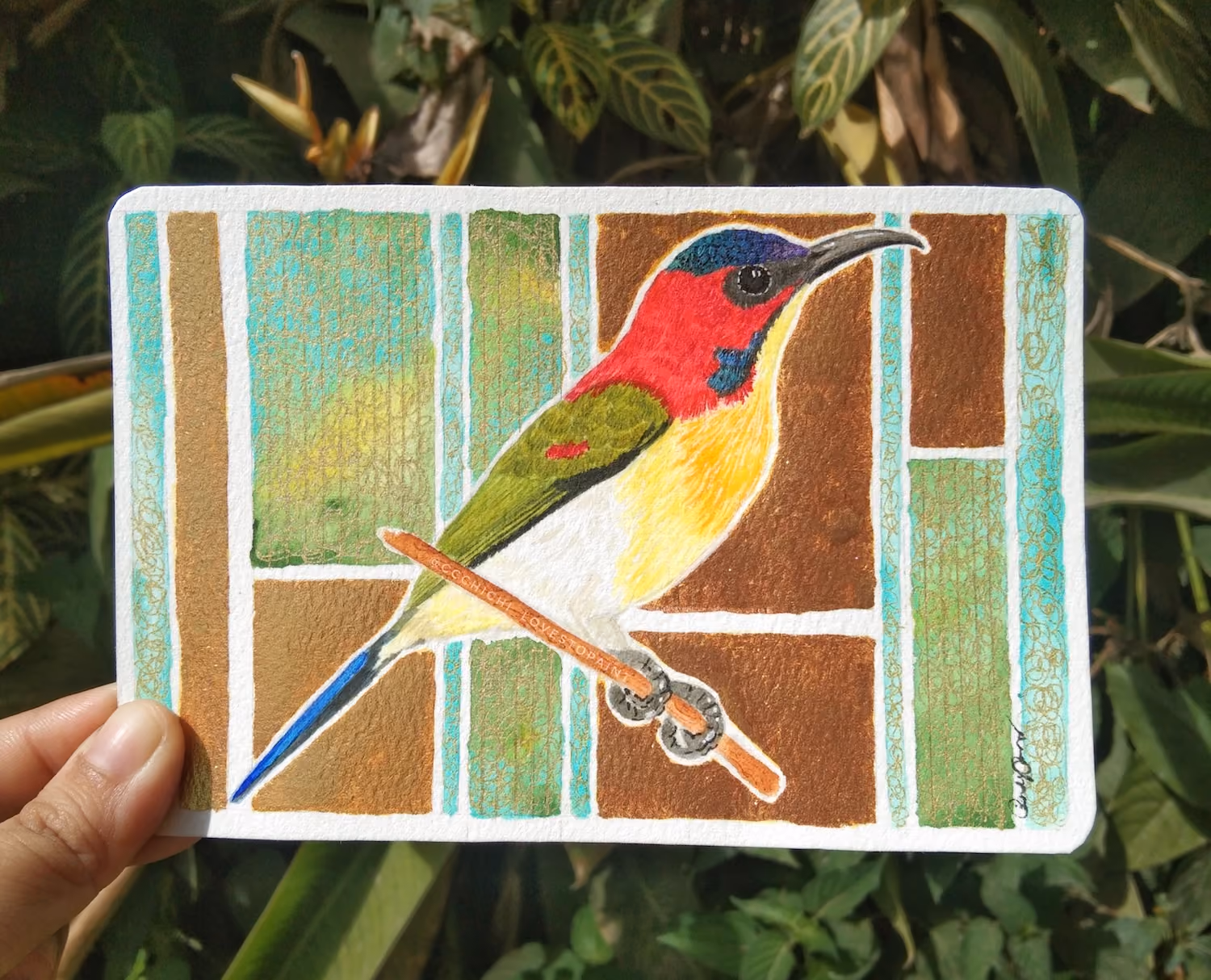 Hand holding a card with a colorful bird drawing on a geometric blue, brown, and green background.