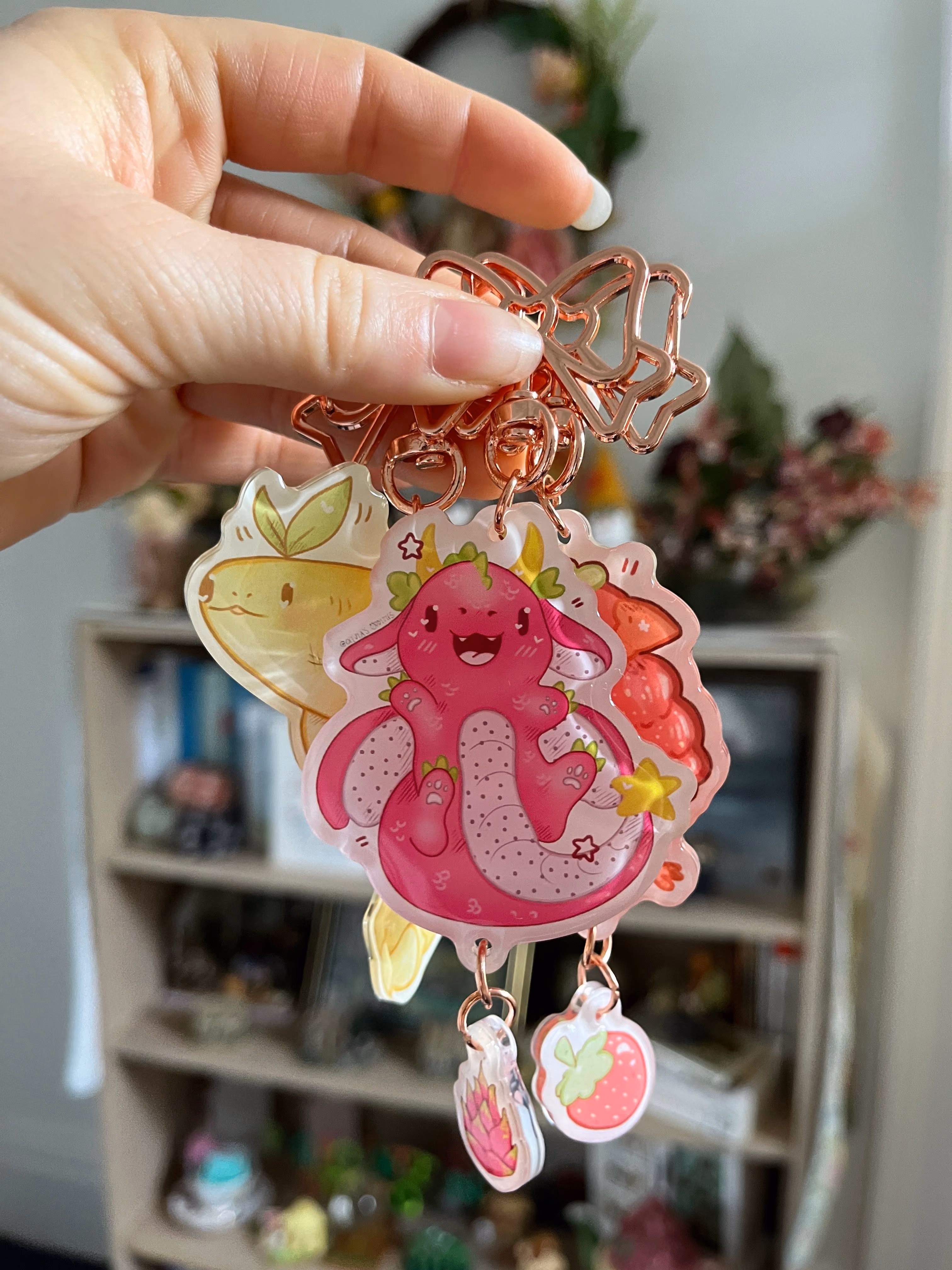 Hand holding keychain with pink axolotl and fruit-themed charms against blurred shelf background.