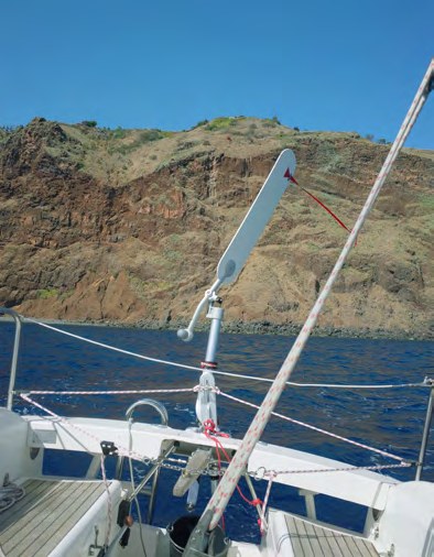 Blick vom Heck des Segelboots auf die Windsteueranlage und eine felsige Küste