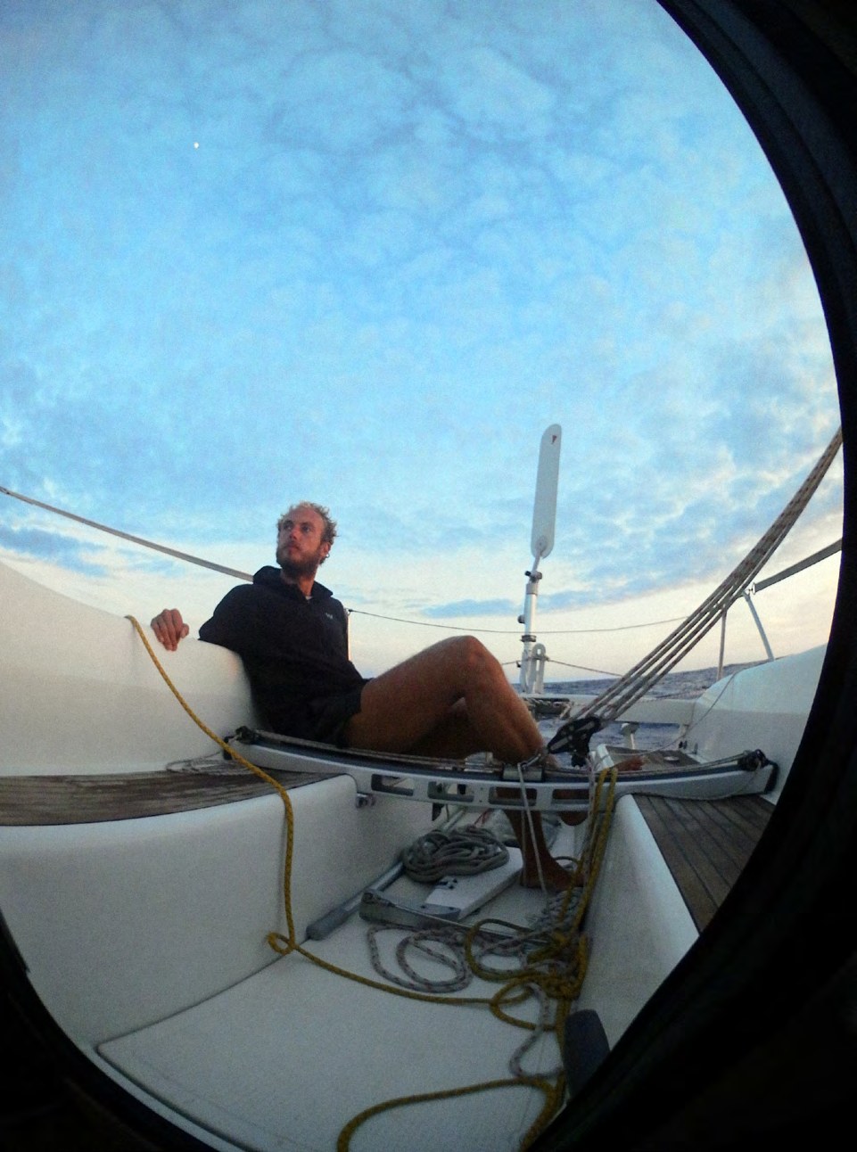 Blick durch ein rundes Bullauge auf Simon, der im Cockpit des Segelboots sitzt
