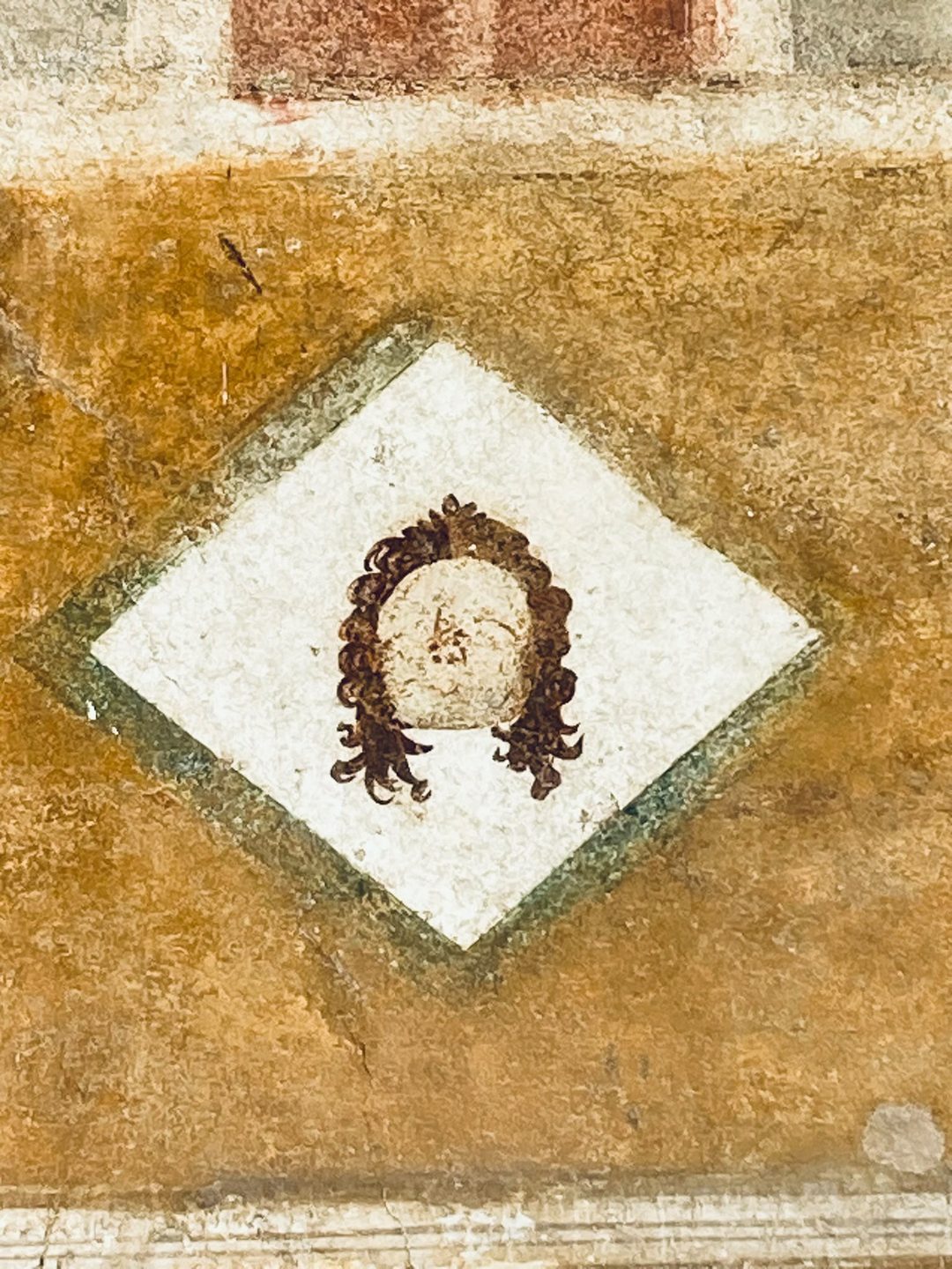Detail einer pompejanischen Wandmalerei: ein gemalter Kopf mit Locken in einer weißen Raute
