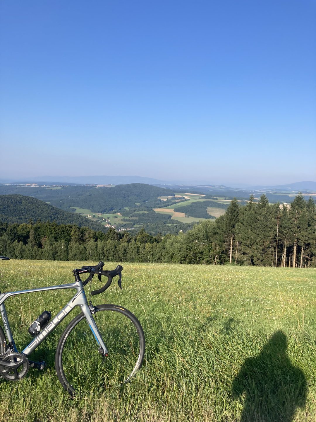 Rennrad in einer Wiese auf einem Hügel, dahinter bewaldete Hügel und weites Land, im Vordergrund der Schatten des Fotografen