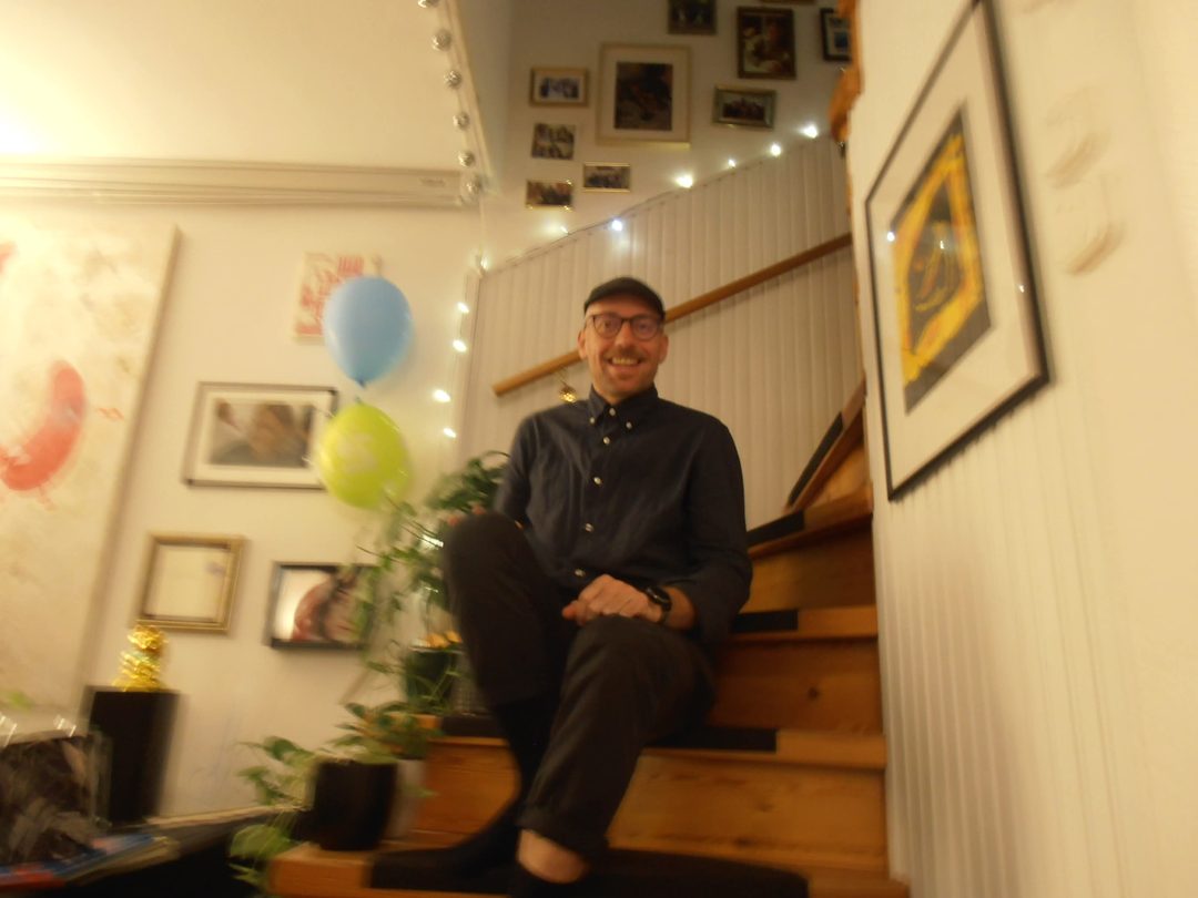 Andreas sitzt lächelnd auf einer Holztreppe, daneben Luftballons und gerahmte Bilder an der Wand