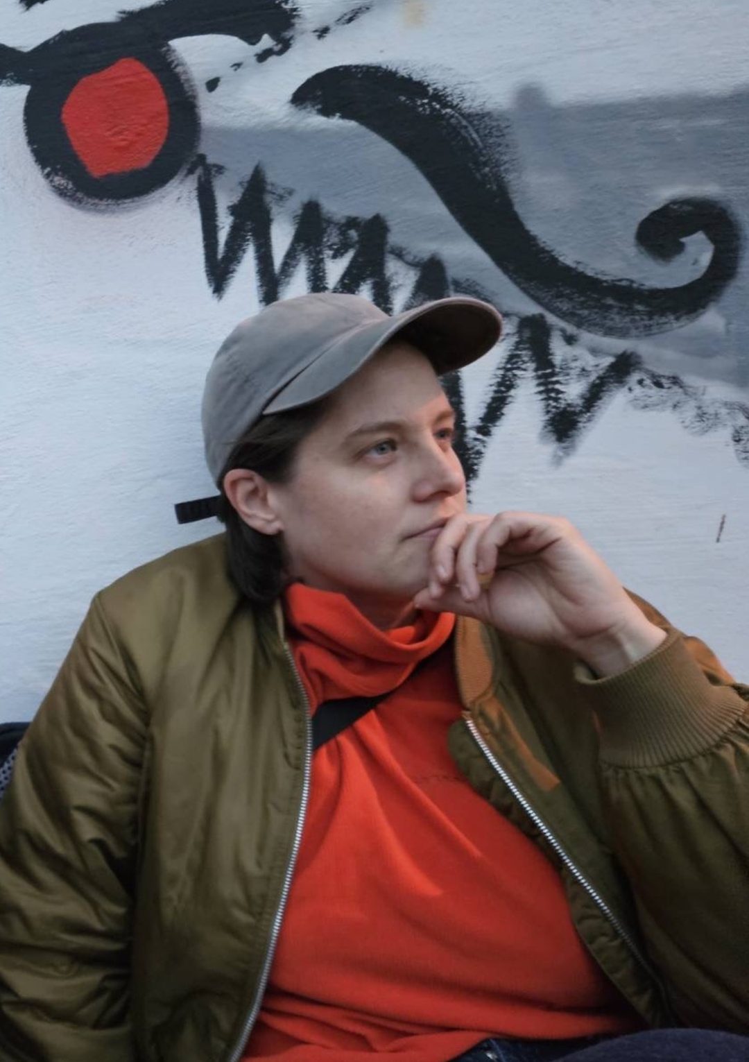 Iris mit grauer Kappe, rotem Rollkragen und Bomberjacke, das Kinn auf die Hand gestützt, vor einer bemalten Wand