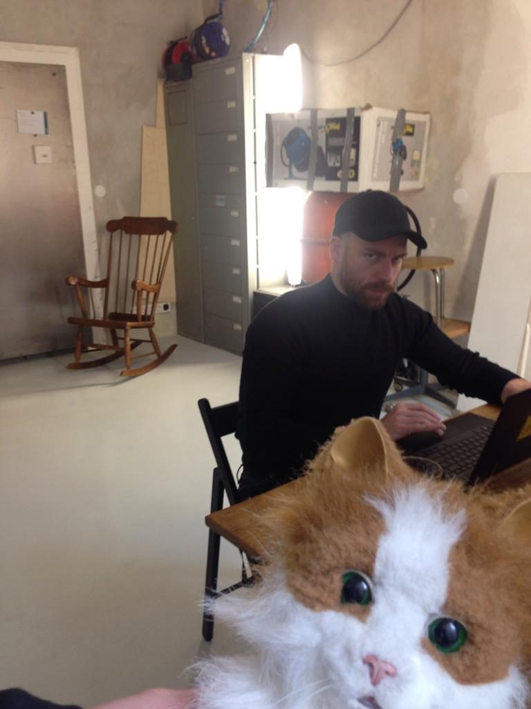 Andreas mit schwarzer Kappe am Laptop in einem Atelier, im Vordergrund eine Katzen-Handpuppe