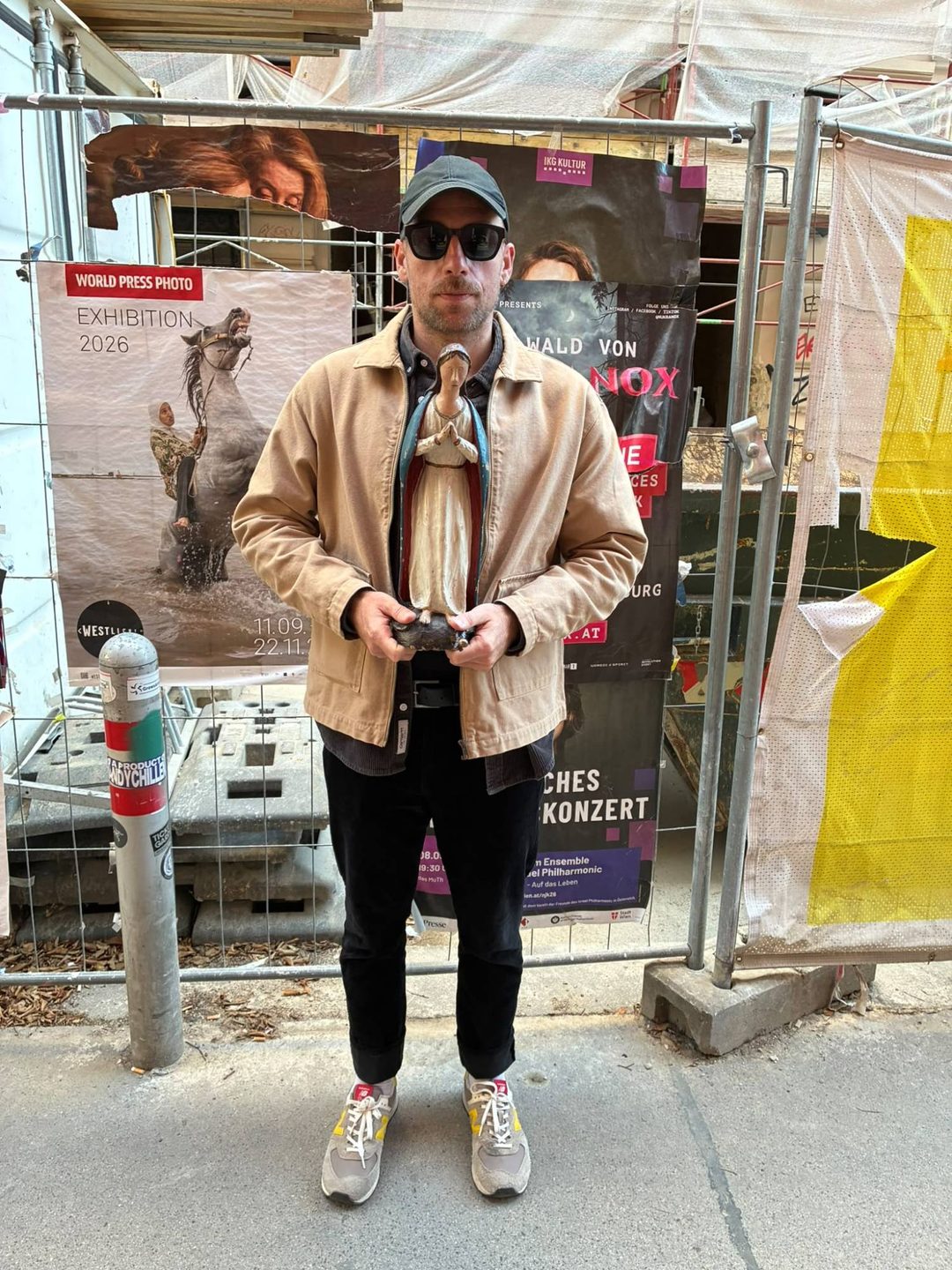 Andreas mit Kappe und Sonnenbrille vor einem Bauzaun mit Plakaten, in den Händen eine bemalte Holzfigur einer betenden Madonna