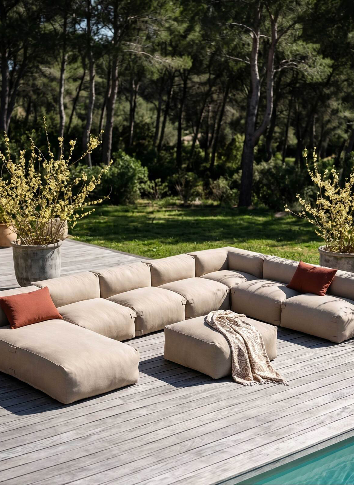 Canapé d'extérieur modulable beige en forme de U avec deux coussins orange, une ottomane centrale recouverte d'un plaid, sur une terrasse en bois près d'une piscine et entourée de verdure et d'arbres.