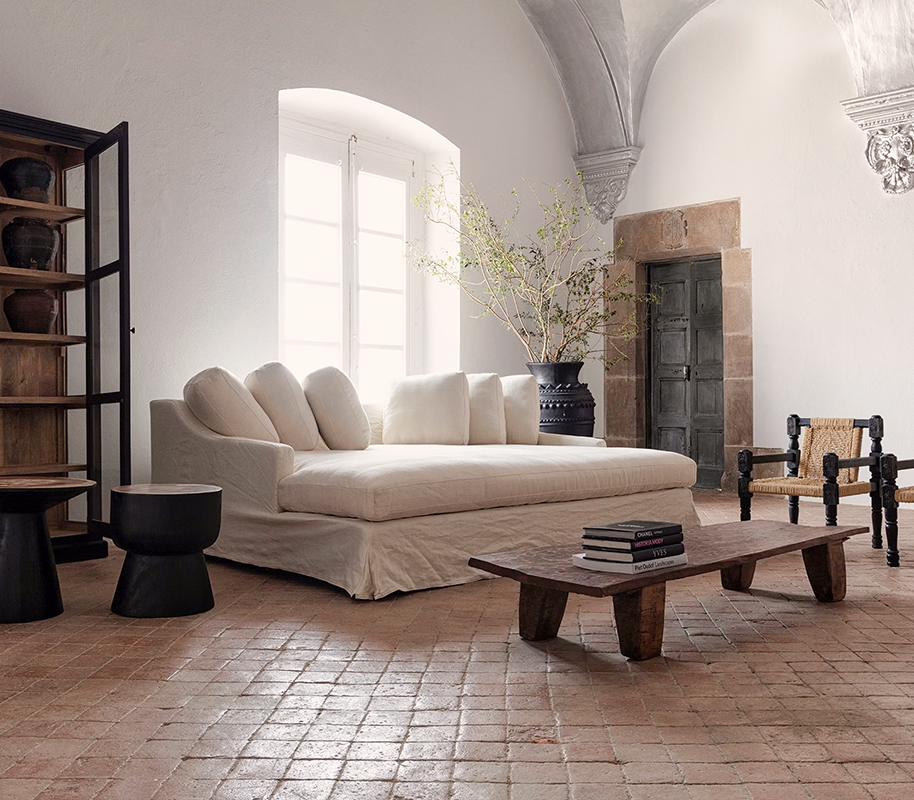 Salon minimaliste avec un canapé blanc moelleux devant une grande fenêtre lumineuse, une table basse en bois avec des livres, une chaise en bois et corde, un grand vase avec des branches et une étagère en bois avec des pots anciens.