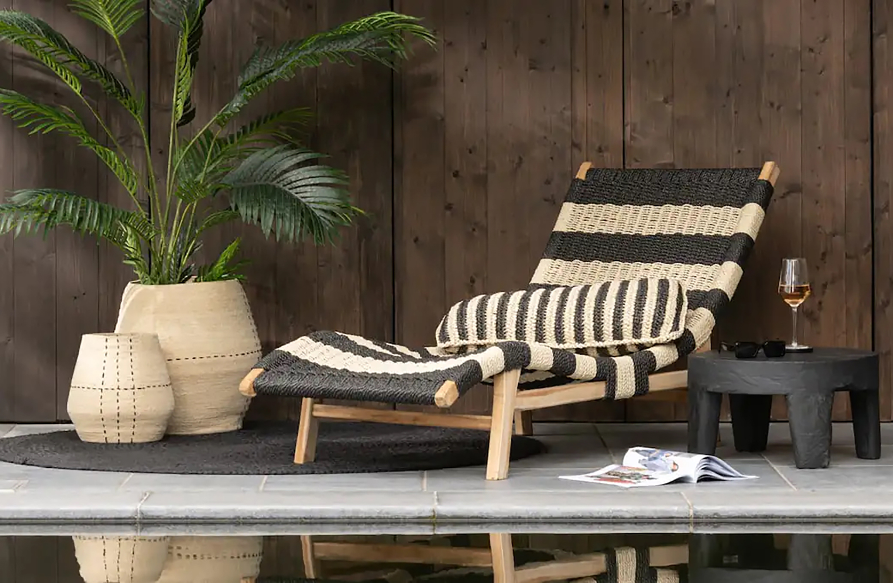 Chaise longue tissée noire et beige avec un coussin rayé assorti, posée sur une terrasse carrelée à côté d'une petite table noire avec un verre de vin blanc et des lunettes, devant un mur en bois foncé, avec deux grands pots en fibres naturelles contenant une plante verte.