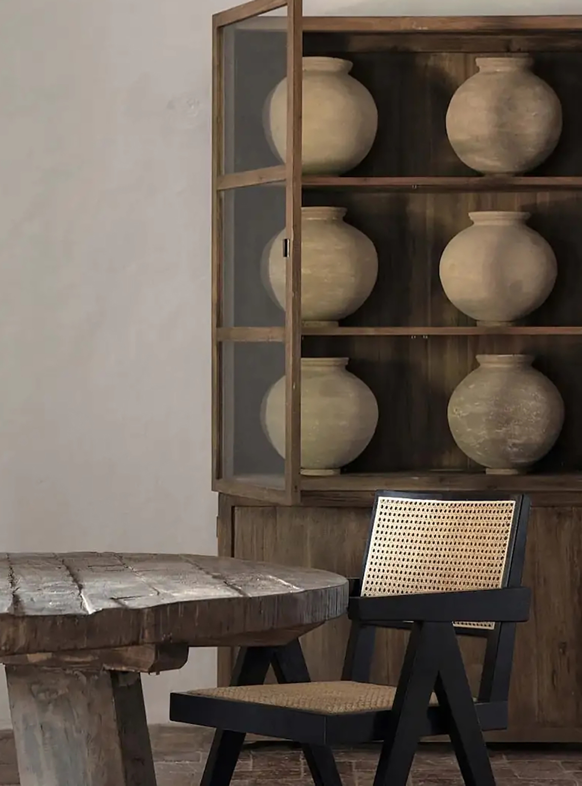 Coin salle à manger avec une table ronde en bois brut, une chaise noire avec dossier tressé beige et un buffet en bois avec portes en verre contenant six pots en terre cuite.