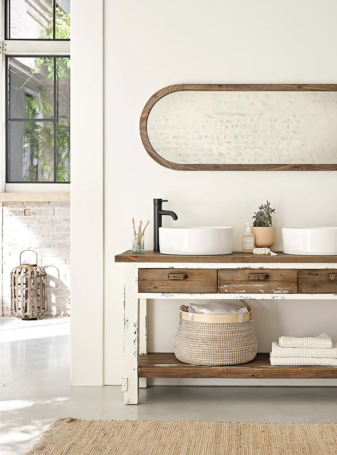 Salle de bain lumineuse au style rustique avec un meuble en bois blanc vieilli, deux vasques blanches ovales, un robinet noir, un miroir ovale encadré en bois, des brosses à dents, un distributeur de savon, une plante en pot, un panier tissé avec serviettes et une fenêtre donnant sur un espace extérieur végétal.