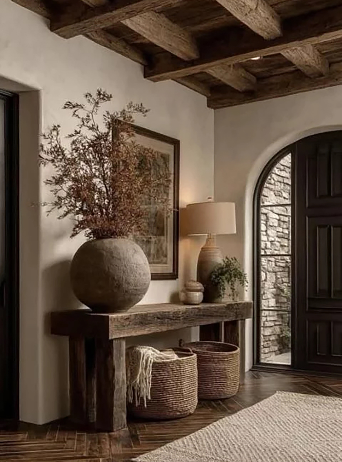 Entrée chaleureuse avec plafond à poutres en bois, console rustique en bois massif surmontée d'un grand vase rond avec branches sèches, une lampe sur pied, une plante verte en pot, et deux paniers tressés sous la console, proche d'une porte d'entrée en bois avec une arche en pierre.