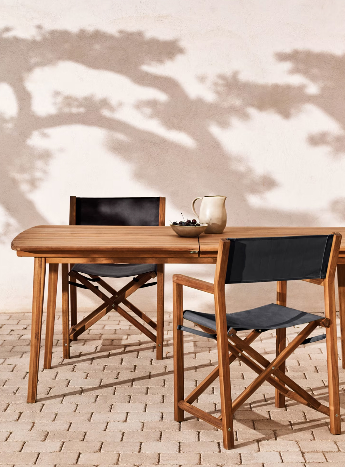Table en bois clair avec deux chaises en bois et toile noire sur un sol pavé, avec une carafe beige et un bol de cerises sur la table et des ombres d'arbres projetées sur un mur clair en arrière-plan.