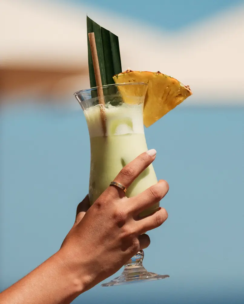 A hand holding a fresh piña colada garnished with a pineapple slice, set against a blue sky and sea — the healthy, fresh beach bar at Casita.