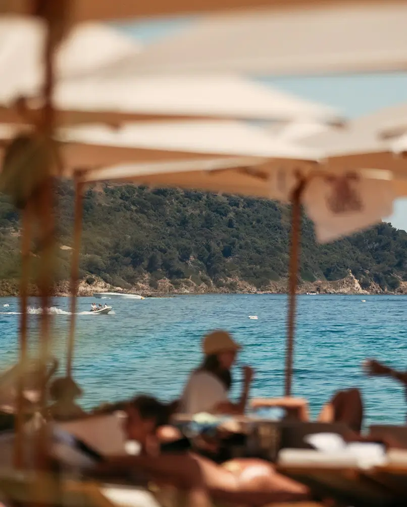 Sunbeds shaded by white parasols look out over the turquoise sea and a green headland, a boat passing by — the relaxing beachside atmosphere at Casita.