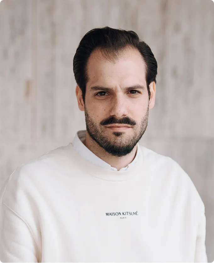 Mann mit dunklem Haar, Bart und weißem Maison Kitsuné Pullover vor verschwommenem Hintergrund.