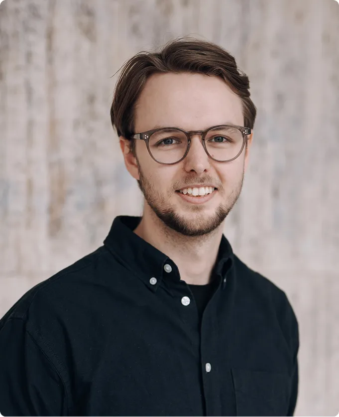 Porträt eines lächelnden jungen Mannes mit Brille in schwarzem Hemd vor unscharfem Hintergrund.