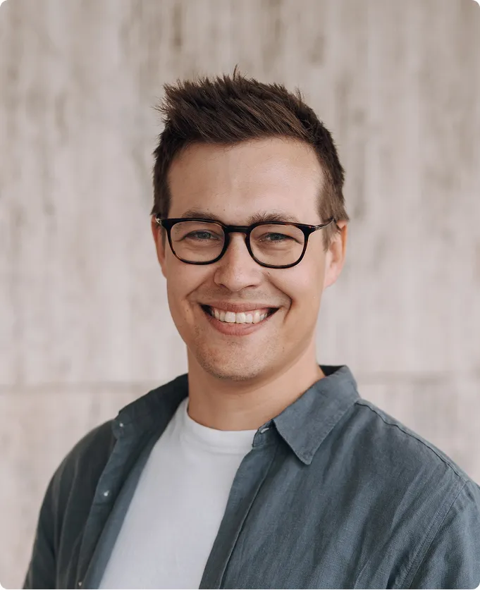 Porträt eines lächelnden Mannes mit Brille, weißem T-Shirt und blauem Hemd vor beigem Hintergrund.