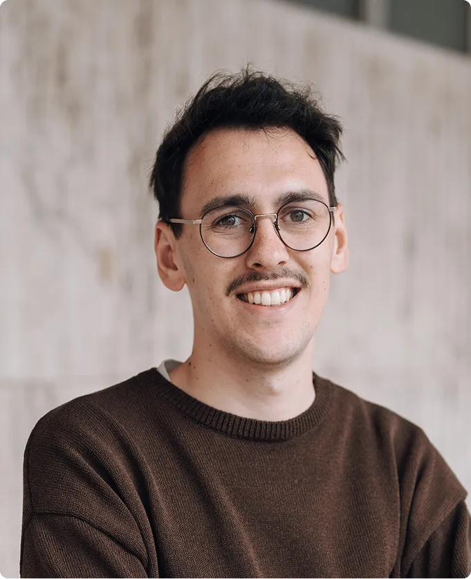 Junger Mann mit Brille und Schnurrbart lächelt, trägt braunen Pullover vor hellem Hintergrund.