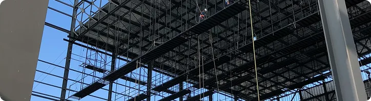 Bauarbeiter steigen auf ein Stahlgerüst in einer Industriehalle bei klarem Himmel.