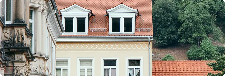 Fassadendetail eines altmodischen Gebäudes mit roten Ziegeldächern und grünen Bäumen im Hintergrund.