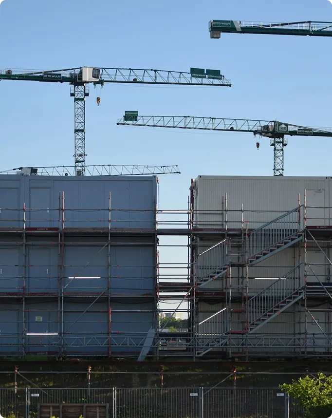 Mehrere Kräne über einer Baustelle mit modularen Gebäuden und Gerüsten unter klarem Himmel.