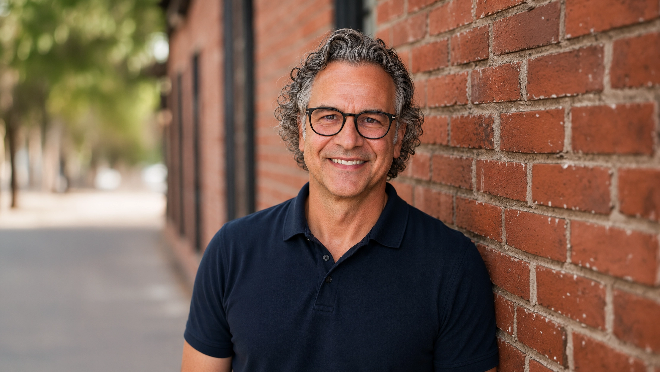 Smiling middle-aged man with curly gray hair and glasses wearing a navy blue polo shirt, leaning against a brick wall outdoors with a blurred street and trees in the background.