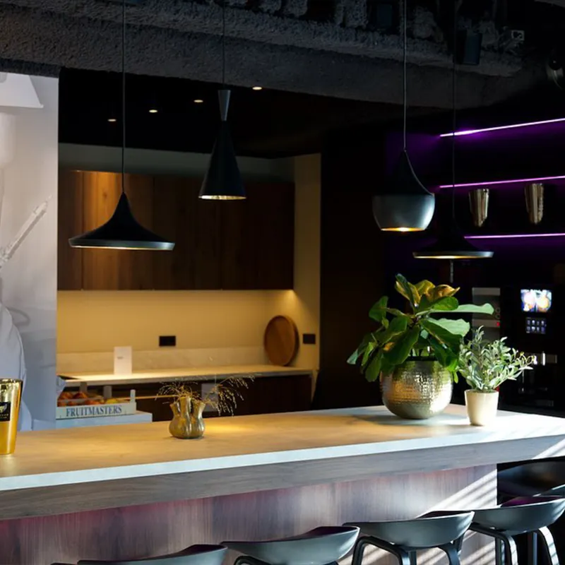 Moderne bar met houten blad, zwarte barkrukken, hanglampen en planten in potten.