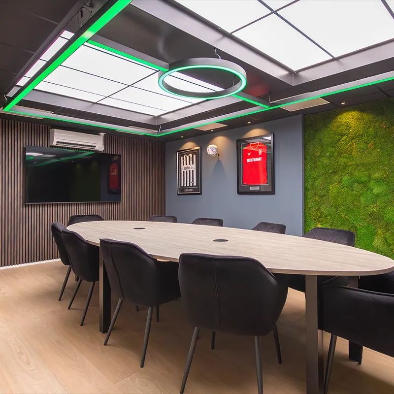 Moderne vergaderruimte met ovale houten tafel, zwarte stoelen, groene mosmuur en ingelijste voetbalshirts.