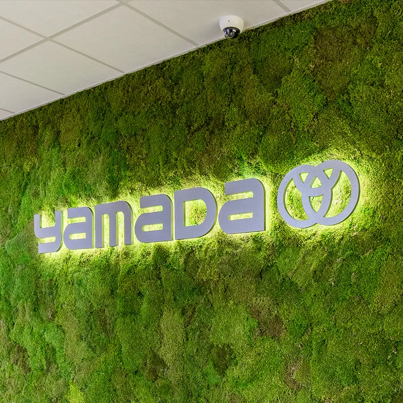 Verlicht logo 'yamada' met een gestileerd symbool op een groene mosmuur.