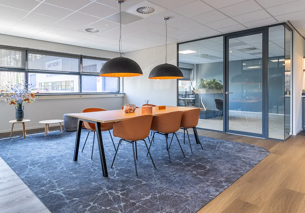 Moderne vergaderruimte met houten tafel, vier oranje stoelen en grote ramen naast glazen wand.