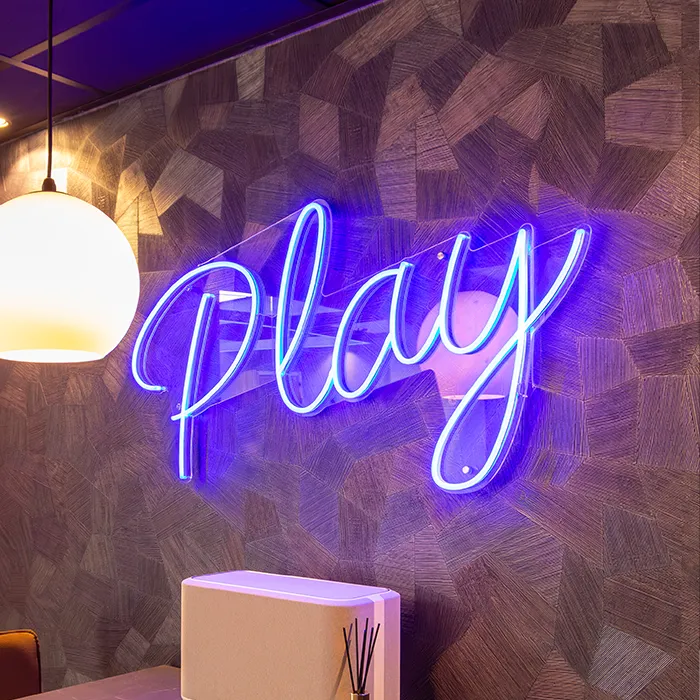 Neonblauw signaal met het woord 'play' aan een muur met een geometrisch patroon en een witte lamp.