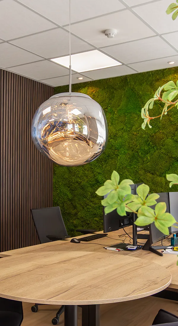 Moderne kantoorruimte met hangende glazen lamp, houten tafels en groene moswand.