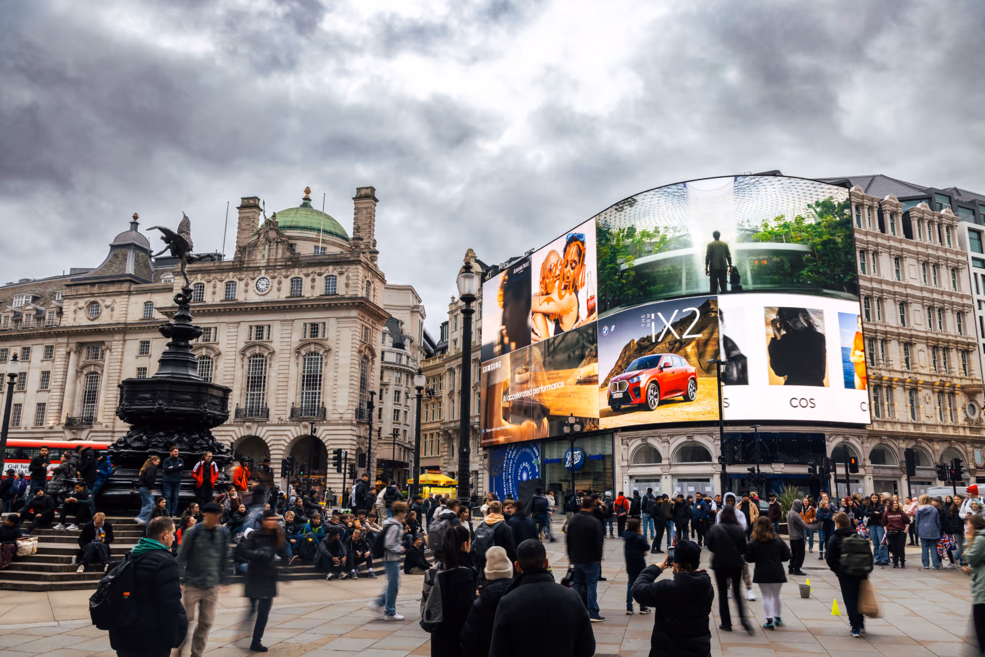 Crowd gathered around the Shaftesbury Memorial Fountain with digital advertisements displayed on large curved screens in London's Piccadilly Circus under a cloudy sky.