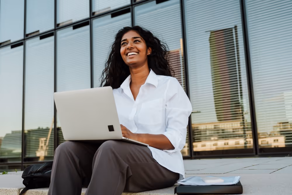 Young person with curly hair focused on a laptop while sitting at a wooden table with an open notebook.