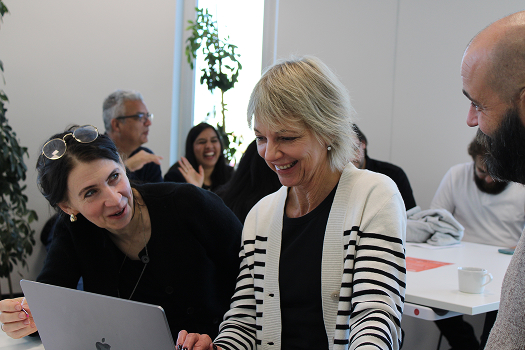 Three coworkers smiling and talking while looking at a laptop in a bright office setting.