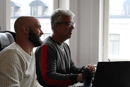 Two men sitting side by side looking at a laptop screen in a room with large windows.