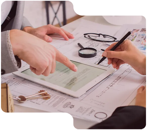 Two people reviewing documents and plans with a tablet, magnifying glass, and glasses on the table.
